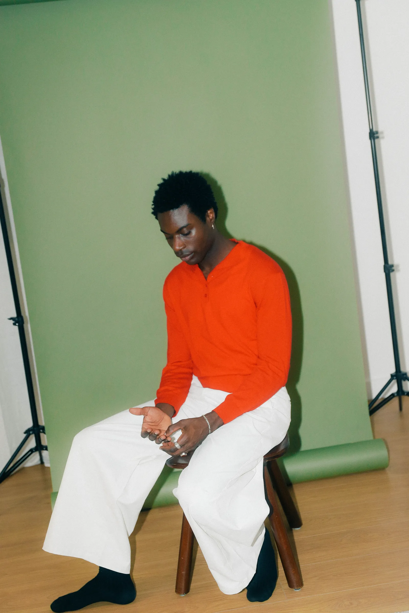 Personne assise sur un tabouret en bois, devant un fond vert, en pull orange et pantalon blanc.