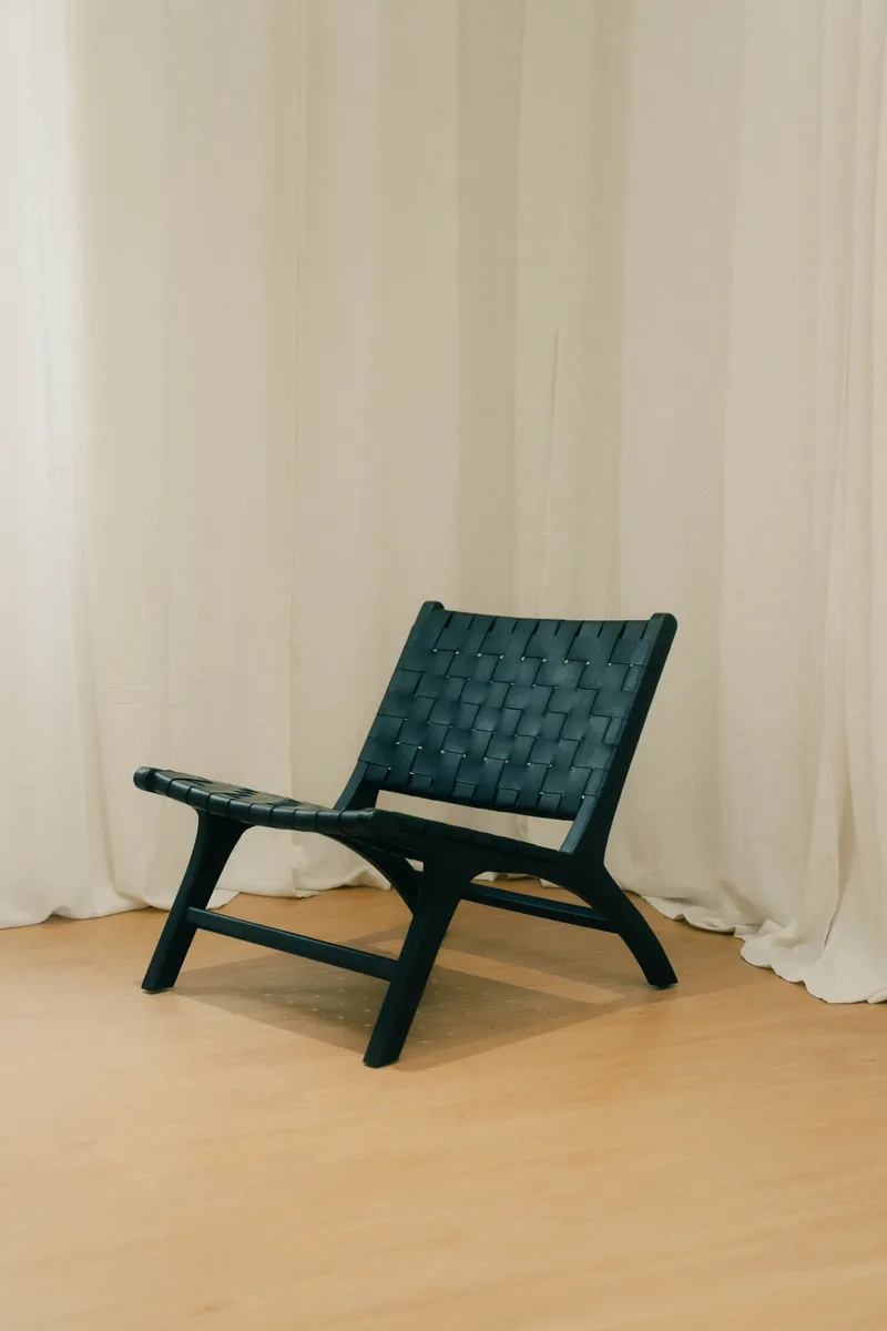 Fauteuil en cuir tressé noir à structure en bois sombre, devant un rideau de lin.